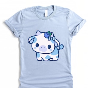 May include: Light blue t-shirt featuring a cartoon cow with white fur and blue spots. The cow has a blueberry and bow on its head. The shirt is made of soft material and has rolled-up sleeves.