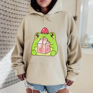 May include: A light beige hoodie featuring a cartoon frog wearing a strawberry on its head and drinking strawberry milk from a carton.