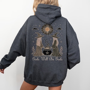 Puede incluir: Sudadera con capucha gris oscuro con un gráfico de dos brujas, un caldero, un sol y el texto "Girls Will Be Girls". El diseño es en tonos marrones y dorados. La sudadera tiene capucha y mangas largas.