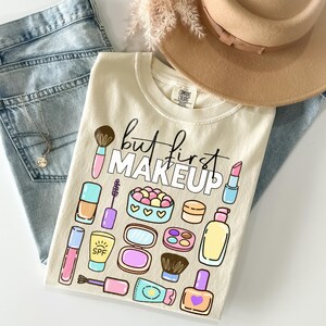 May include: Cream t-shirt featuring the text "but first MAKEUP" and makeup illustrations. The shirt is styled with light blue jeans, a gold necklace, and a tan hat. Makeup illustrations include a brush, lipstick, and nail polish.