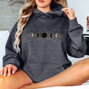 Puede incluir: Una sudadera con capucha gris oscuro con un dise&ntilde;o de fases lunares doradas. El dise&ntilde;o presenta una luna creciente, una media luna, una luna llena y una luna gibosa menguante.