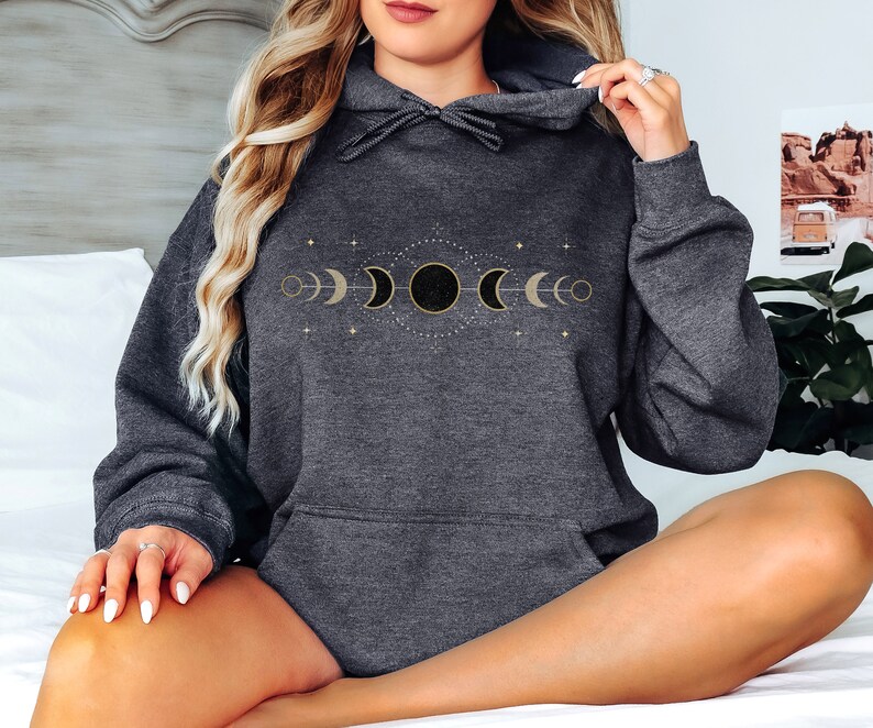 Puede incluir: Una sudadera con capucha gris oscuro con un dise&ntilde;o de fases lunares doradas. El dise&ntilde;o presenta una luna creciente, una media luna, una luna llena y una luna gibosa menguante.