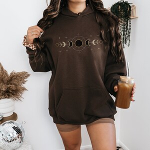 Puede incluir: Una sudadera con capucha marr&oacute;n de gran tama&ntilde;o con un dise&ntilde;o de fases lunares doradas en la parte delantera. La sudadera tiene una capucha con cord&oacute;n y un bolsillo canguro.