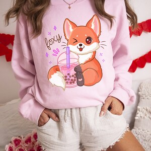 May include: Pink sweatshirt with a cartoon fox holding a boba tea cup. The sweatshirt has the word "foxy" printed on it in purple.