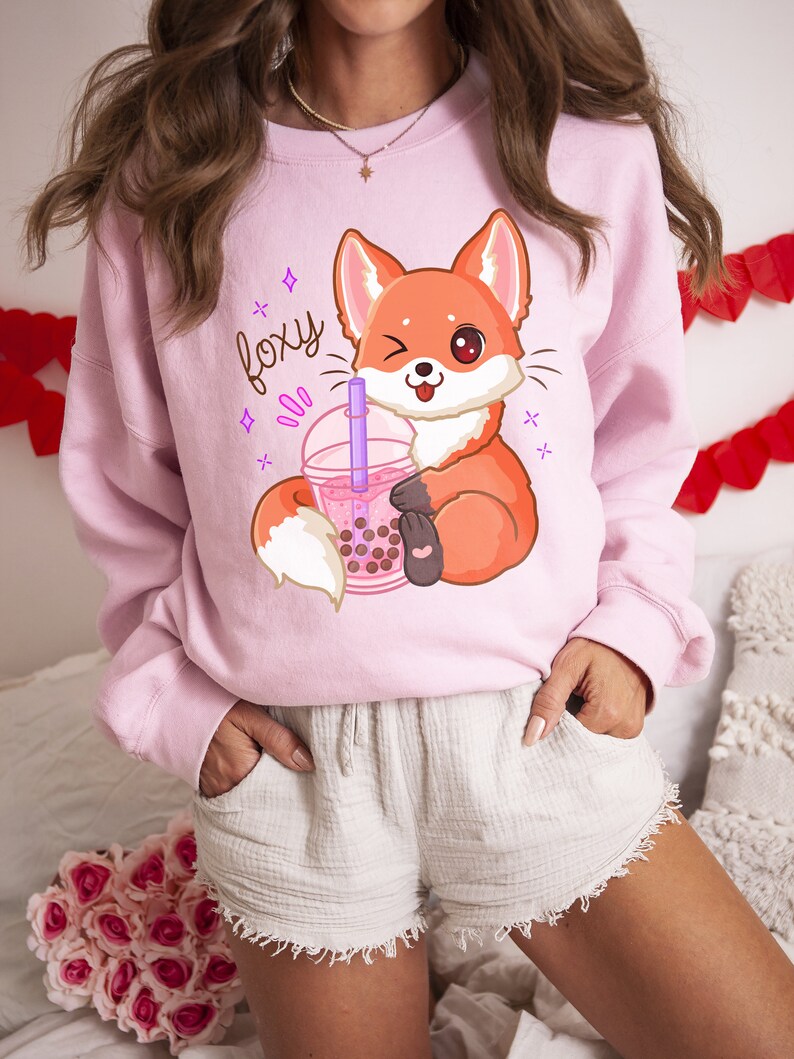 May include: Pink sweatshirt with a cartoon fox holding a boba tea cup. The sweatshirt has the word "foxy" printed on it in purple.
