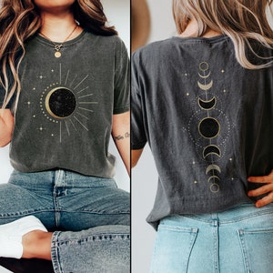 Op de afbeelding: Twee grijze T-shirts met gouden hemelse ontwerpen. Een shirt heeft een zon en maan met sterren, de andere heeft een verticale lijn van manen en sterren.