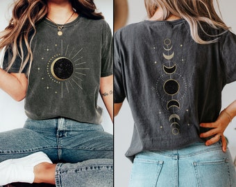 Puede incluir: Dos camisetas grises con diseños celestiales dorados. Una camiseta presenta un sol y una luna con estrellas, la otra presenta una línea vertical de lunas y estrellas.