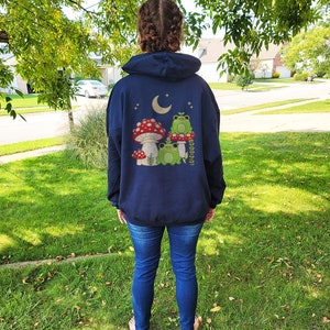 Pu&ograve; includere: Una felpa con cappuccio blu navy con una grafica di tre rane sedute su funghi sotto una luna crescente. La grafica &egrave; in tonalit&agrave; di verde, rosso e bianco.