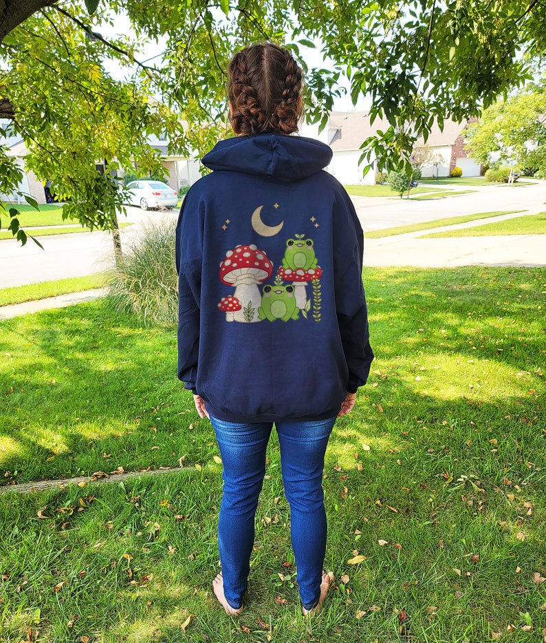 Pu&ograve; includere: Una felpa con cappuccio blu navy con una grafica di tre rane sedute su funghi sotto una luna crescente. La grafica &egrave; in tonalit&agrave; di verde, rosso e bianco.