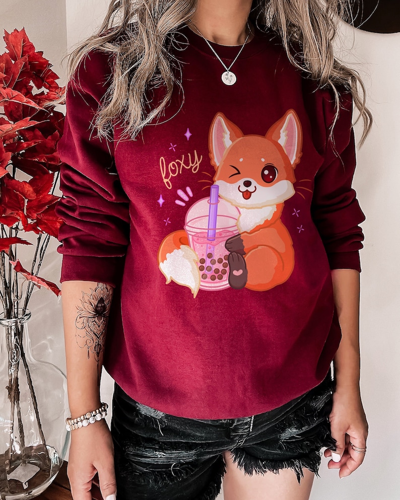 May include: A maroon sweatshirt with a cartoon fox holding a pink boba tea cup. The fox is winking and has a pink nose. The text "foxy" is written in pink above the fox.