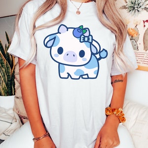 May include: White t-shirt featuring a cartoon cow with a blueberry theme. The cow is white with blue spots and accents, including a blueberry bow. The design is centered on the shirt, creating a playful and whimsical aesthetic.
