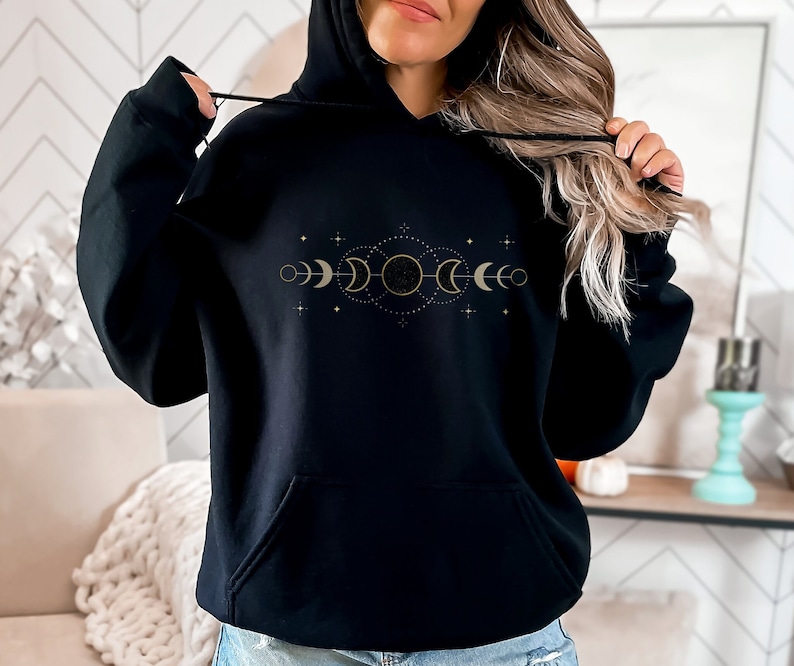 Puede incluir: Sudadera con capucha negra con un dise&ntilde;o de fases lunares doradas. El dise&ntilde;o presenta una luna creciente, luna llena y sol con estrellas.