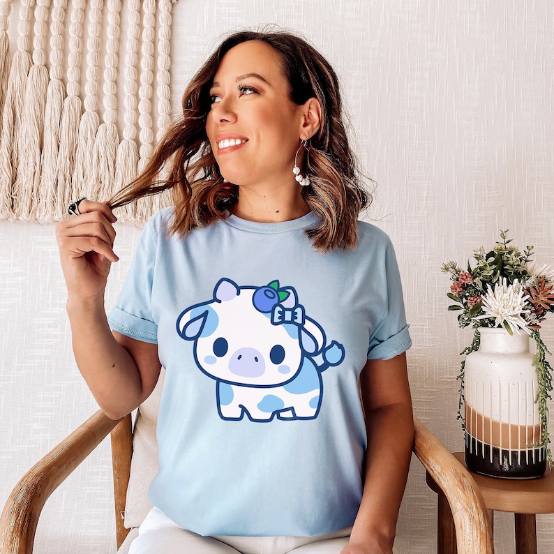 以下が含まれることがあります： ブルーのTシャツに、ブルーベリーのヘッドバンドをした漫画の牛が描かれています。牛は白地に青い斑点があり、かわいい表情をしています。