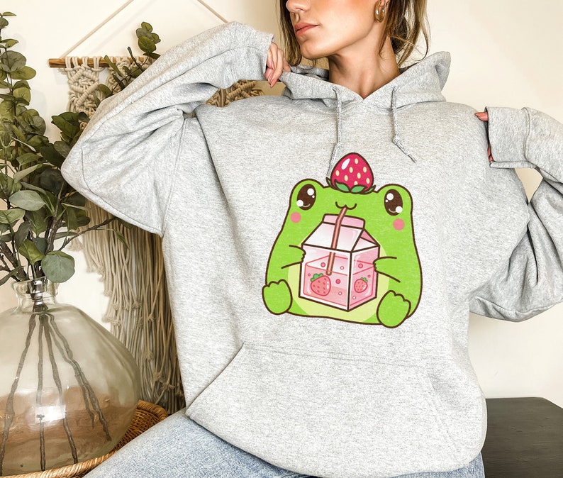 May include: A grey hoodie featuring a cartoon frog wearing a strawberry on its head and drinking strawberry milk from a carton.