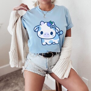May include: Light blue t-shirt featuring a cartoon cow with blue spots and a blueberry bow. The cow has large eyes and a cute expression. The shirt is a soft, casual style, perfect for everyday wear.