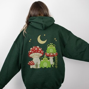 Pu&ograve; includere: Una felpa con cappuccio verde scuro con una stampa di tre rane sedute su funghi sotto una luna crescente. Le rane sono verdi con occhi bianchi e i funghi sono rossi con macchie bianche.