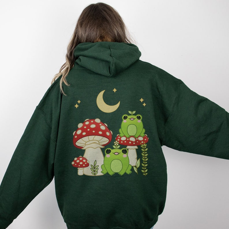 Pu&ograve; includere: Una felpa con cappuccio verde scuro con una stampa di tre rane sedute su funghi sotto una luna crescente. Le rane sono verdi con occhi bianchi e i funghi sono rossi con macchie bianche.
