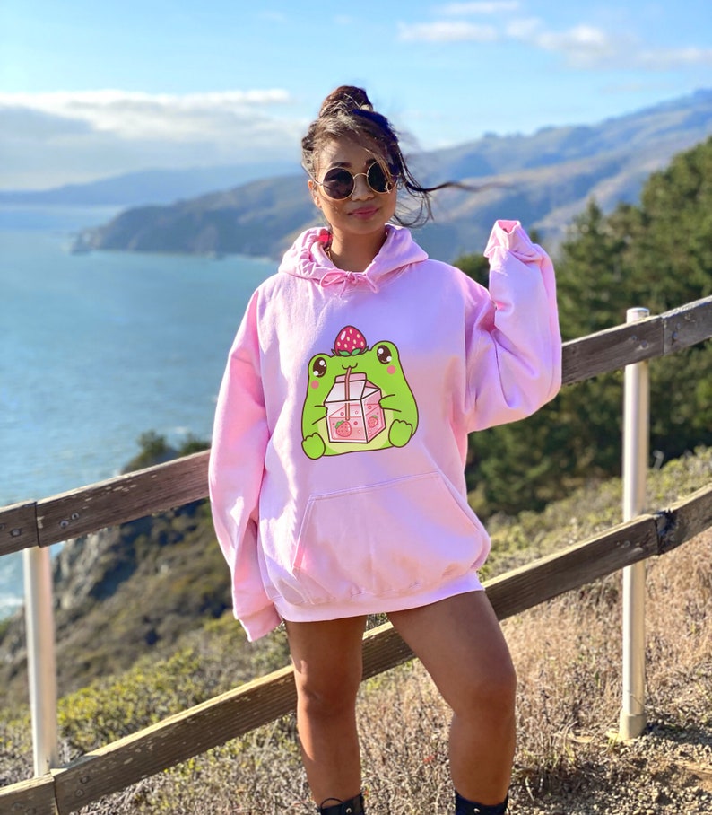 May include: A pink oversized hoodie featuring a cartoon frog drinking strawberry milk. The frog is green with a strawberry on its head.