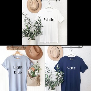 May include: Three t-shirts in white, light blue, and navy blue are displayed on hangers. Each shirt has a word printed on it: "White", "Light Blue", and "Navy". The shirts are hanging on a wall with hats and greenery.