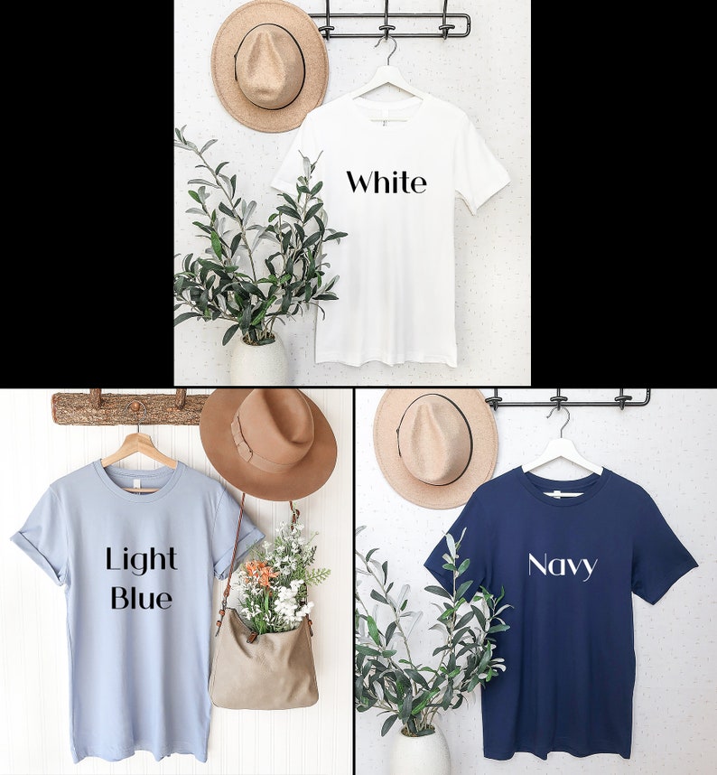 May include: Three t-shirts in white, light blue, and navy blue are displayed on hangers. Each shirt has a word printed on it: "White", "Light Blue", and "Navy". The shirts are hanging on a wall with hats and greenery.