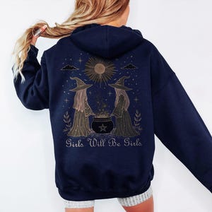 Pu&ograve; includere: Felpa con cappuccio blu navy con una grafica sul retro raffigurante due streghe, un calderone e la scritta "Girls Will Be Girls". Il design include un sole, stelle ed elementi decorativi. La felpa ha un cappuccio e maniche lunghe.