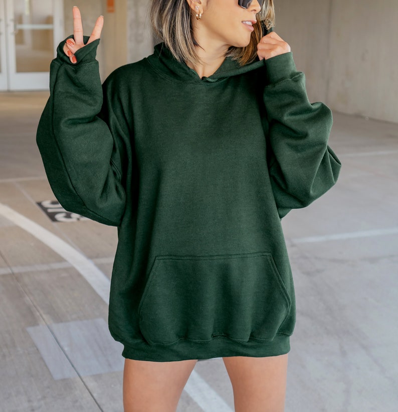 Pu&ograve; includere: Una felpa con cappuccio oversize di colore verde scuro con una tasca a marsupio. La felpa ha un cappuccio con coulisse.
