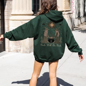 Pu&ograve; includere: Felpa con cappuccio verde scuro con una grafica sul retro raffigurante due streghe, un calderone e la scritta "Girls Will Be Girls". Il design include un sole e delle stelle. La felpa ha un cappuccio con coulisse e maniche lunghe.