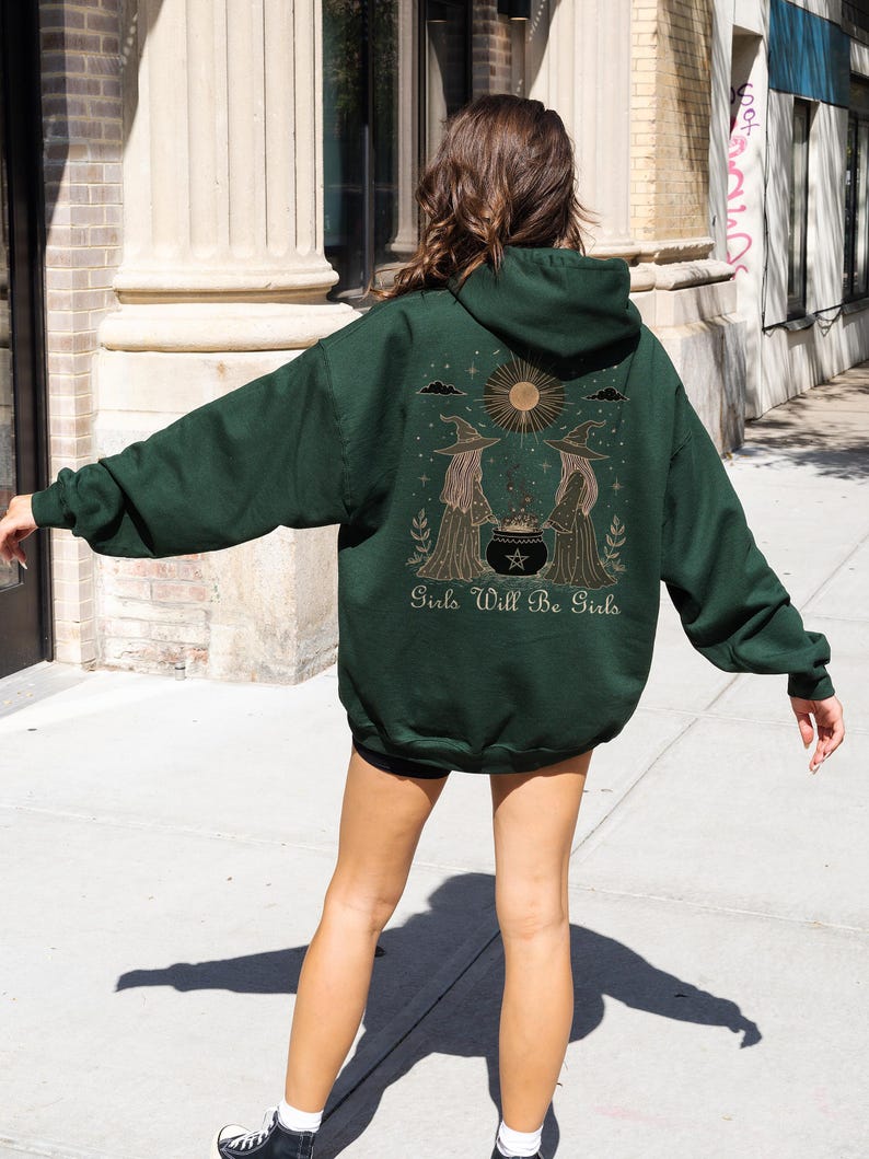 Pu&ograve; includere: Felpa con cappuccio verde scuro con una grafica sul retro raffigurante due streghe, un calderone e la scritta "Girls Will Be Girls". Il design include un sole e delle stelle. La felpa ha un cappuccio con coulisse e maniche lunghe.