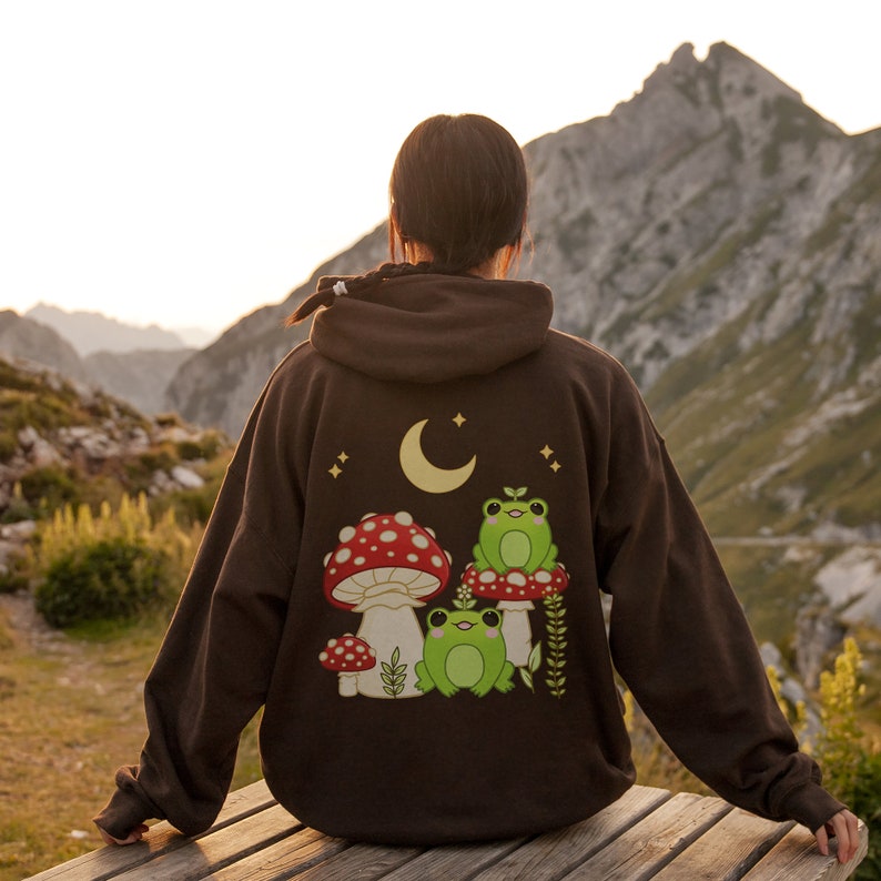 Pu&ograve; includere: Una felpa con cappuccio marrone con una grafica di una luna crescente, stelle, funghi e rane. La felpa ha un cappuccio con coulisse e una tasca a marsupio.