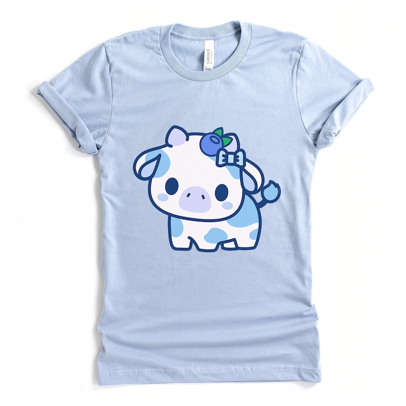May include: Light blue t-shirt featuring a cartoon cow with white fur and blue spots. The cow has a blueberry and bow on its head. The shirt is made of soft material and has rolled-up sleeves.