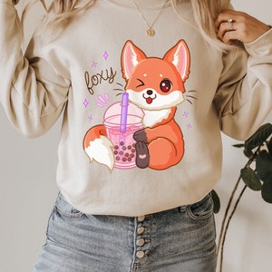 May include: A light beige sweatshirt with a cartoon fox holding a pink boba tea cup. The text "foxy" is written in purple above the fox.