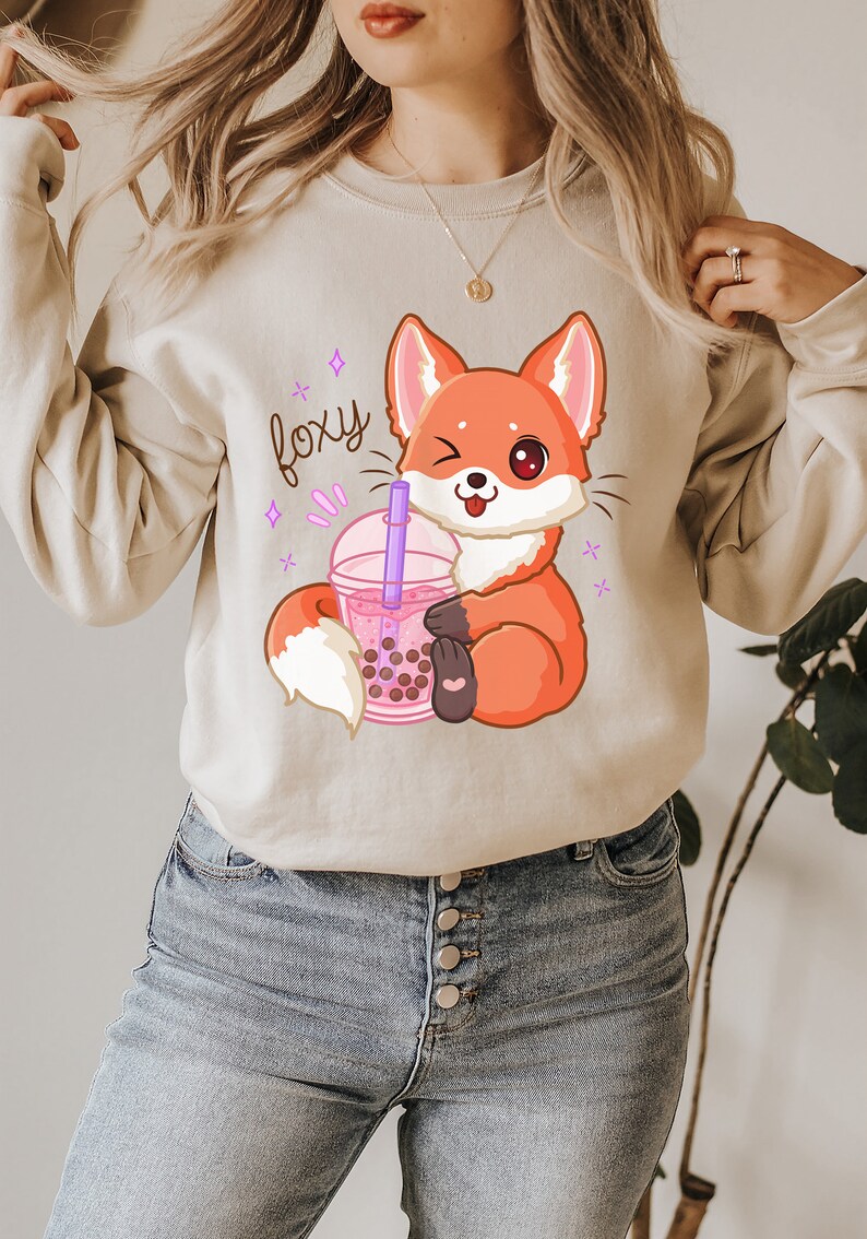 May include: A light beige sweatshirt with a cartoon fox holding a pink boba tea cup. The text "foxy" is written in purple above the fox.