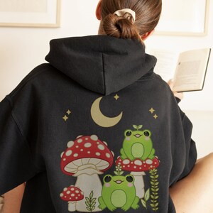 Pu&ograve; includere: Felpa con cappuccio nera con una stampa grafica di tre rane verdi sedute su funghi rossi e bianchi con una luna crescente e stelle sullo sfondo.
