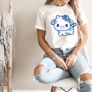 May include: White t-shirt featuring a cartoon cow with blue spots and a blueberry bow. The cow has a cute, friendly expression. The shirt is worn by a person in ripped blue jeans.