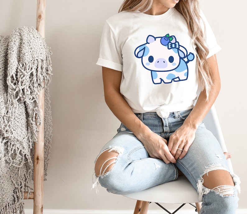 May include: White t-shirt featuring a cartoon cow with blue spots and a blueberry bow. The cow has a cute, friendly expression. The shirt is worn by a person in ripped blue jeans.