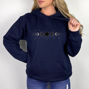 Puede incluir: Una sudadera con capucha azul marino con un dise&ntilde;o de fases lunares doradas en la parte delantera. El dise&ntilde;o presenta una luna creciente, una luna llena y una luna nueva.