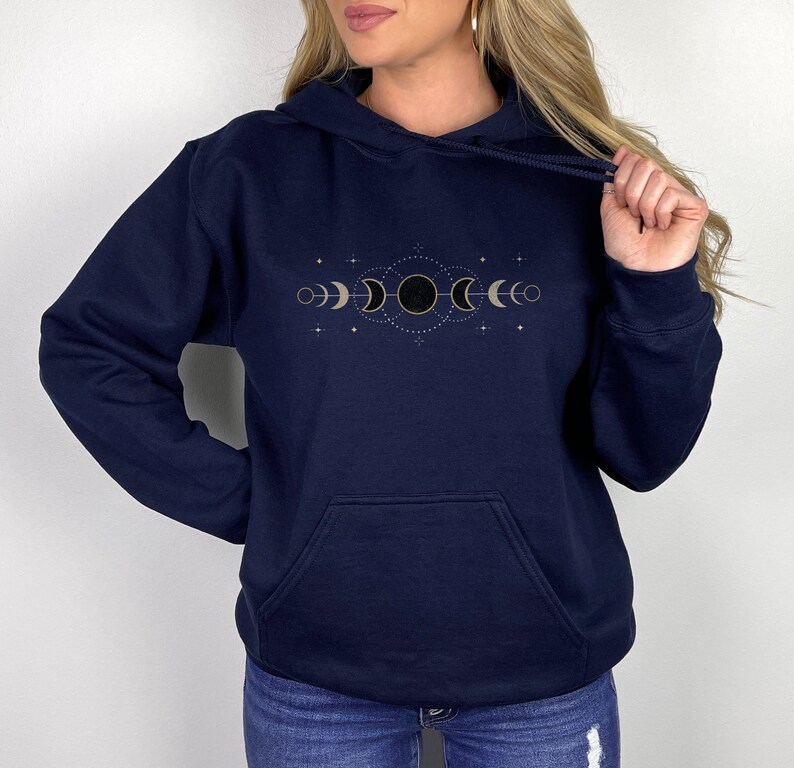 Puede incluir: Una sudadera con capucha azul marino con un dise&ntilde;o de fases lunares doradas en la parte delantera. El dise&ntilde;o presenta una luna creciente, una luna llena y una luna nueva.