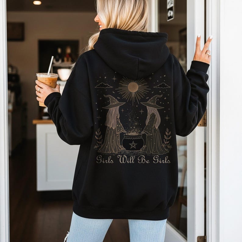 Pu&ograve; includere: Felpa con cappuccio nera con un'illustrazione dorata sul retro raffigurante due streghe, un calderone e la scritta "Girls Will Be Girls". Il design include un sole, stelle e nuvole. La felpa ha un cappuccio con coulisse e una tasca frontale.