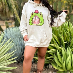 May include: A cream-coloured hoodie featuring a cartoon frog wearing a strawberry on its head and drinking from a carton of strawberry milk. The hoodie has a drawstring hood and a kangaroo pocket.