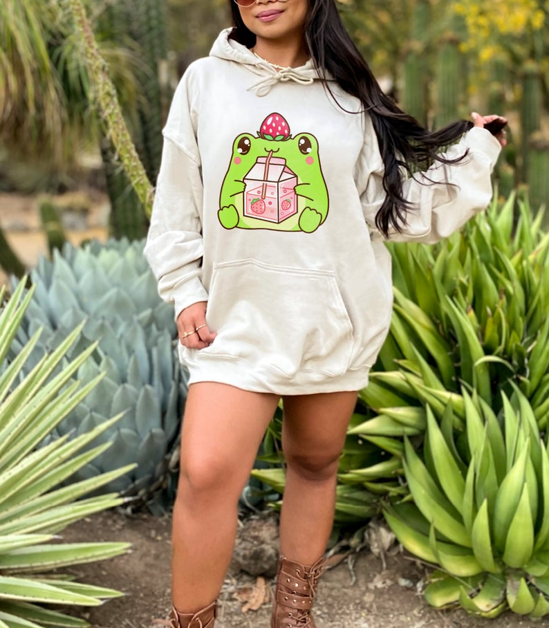 May include: A cream-coloured hoodie featuring a cartoon frog wearing a strawberry on its head and drinking from a carton of strawberry milk. The hoodie has a drawstring hood and a kangaroo pocket.