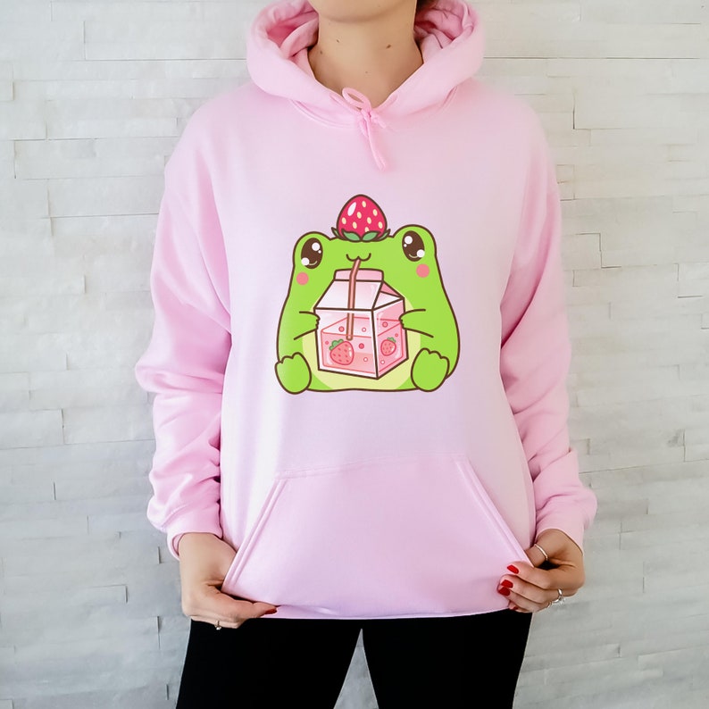 May include: A pink hoodie featuring a cartoon frog wearing a strawberry on its head and drinking strawberry milk from a carton.