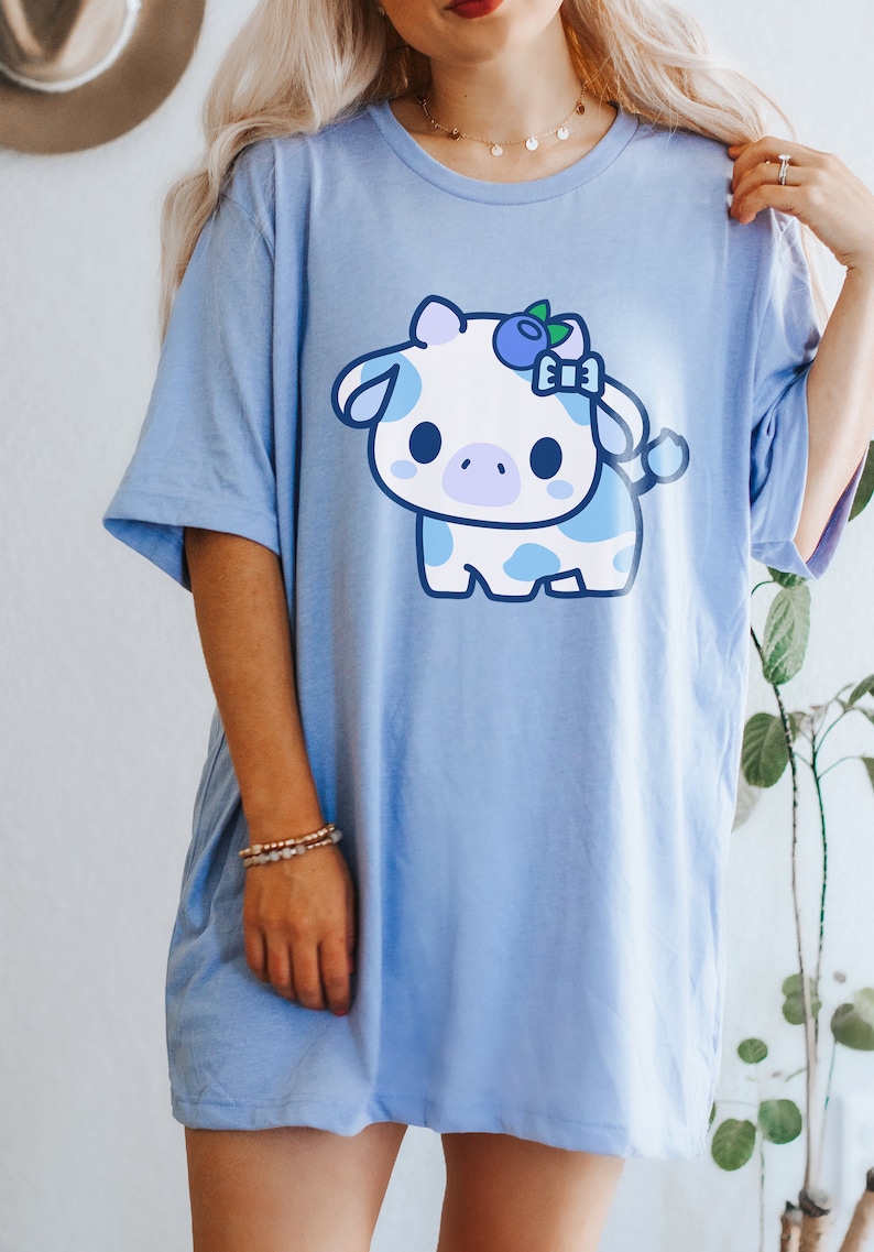 May include: A light blue t-shirt featuring a cartoon cow with a blueberry bow. The cow is white with blue spots and a light purple nose. The shirt has short sleeves and a relaxed fit.