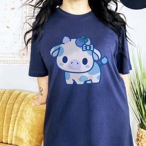 May include: Navy blue t-shirt featuring a cute cartoon cow with blue and white spots, a blueberry bow, and a blueberry on its head. The cow design is centered on the front of the shirt. The shirt is made of a soft, comfortable material.