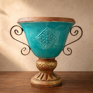 May include: A turquoise ceramic urn with a decorative diamond pattern and ornate gold base. The urn has dark metal handles and a brown rim. The decorative vase is a stylish home decor accent.