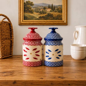 May include: Two ceramic canisters with lids, one featuring red floral patterns and the other featuring blue floral patterns on a cream background. Both canisters have a cylindrical body, a matching patterned lid with a knob, and a painted design.