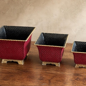 May include: Three decorative square planters in varying sizes. The planters are a deep red with a textured pattern, black interiors, and gold-colored trim and feet. They are arranged on a wooden surface.