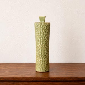 May include: A tall, cylindrical ceramic vase in a light sage green color. The exterior surface features a consistent, dimpled, and textured pattern throughout the body. The vase has a flat top with a small, square-shaped central opening or stopper.