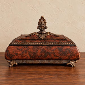 May include: A rectangular box with a dark reddish-brown mottled finish and black accents. The lid features a central decorative finial with leaf-like detailing. The base is supported by four small, ornate feet with matching dark metal trim.