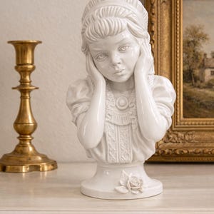 May include: A white glazed ceramic bust of a young person with hands held to their cheeks. The figure wears a ruffled top with a central circular pendant and a headband. A single sculpted rose is attached to the base of the pedestal.