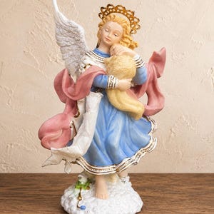 May include: A figurine of an angel with blonde hair, a gold halo, and white wings, wearing a blue dress with a pink sash. The angel holds a small lamb in its arms and stands on a white cloud base with a small green toy sheep at its feet.